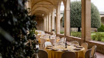 Un espace de restauration en plein air avec des tables rondes dressées pour un repas, entouré d’arcades et de verdure.