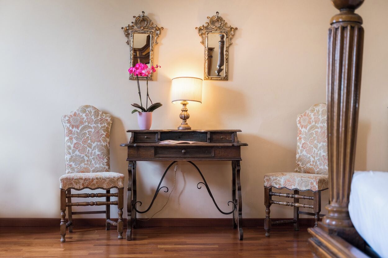 A room with a wooden desk, two chairs, a lamp, and two ornate mirrors on the wall.