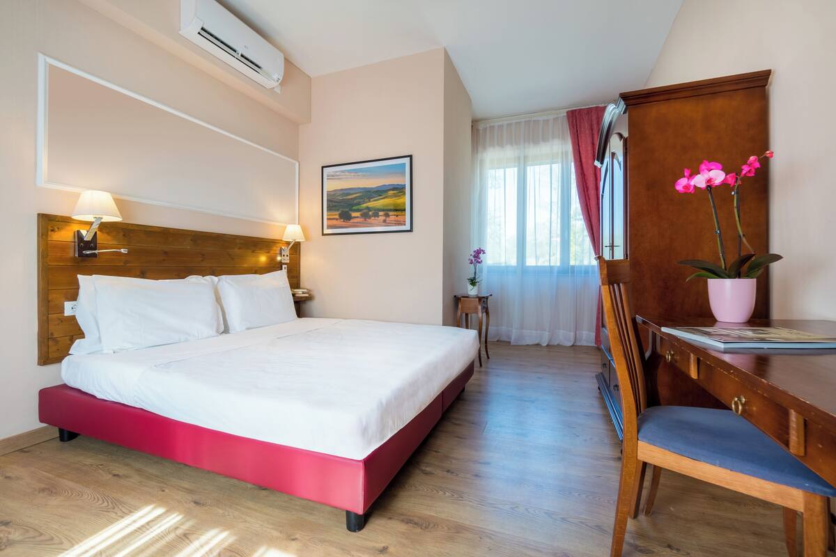 Habitación de hotel con cama, escritorio, silla, mesita pequeña, un cuadro en la pared y ventana con cortinas.