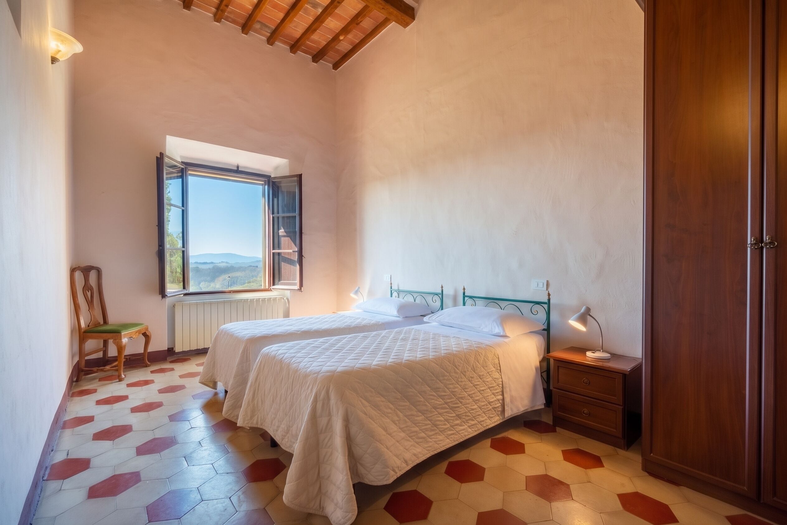 Un dormitorio con dos camas, una ventana con vistas, una silla y una mesita de noche con lámpara.