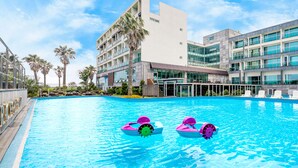 A swimming pool with two inflatable toys, surrounded by modern buildings and palm trees. - The Four Graces Resort (Seogwipo)
