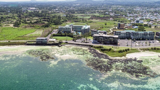 An aerial view of a coastal area with modern buildings, clear water, and surrounding greenery. - The Four Graces Resort (Seogwipo)