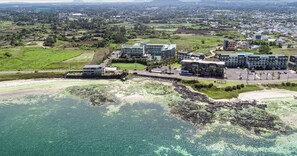 An aerial view of a coastal area with modern buildings, clear water, and surrounding greenery. - The Four Graces Resort (Seogwipo)