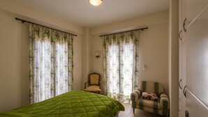 A bedroom with a green quilted bed, two chairs, floral curtains, and a ceiling light. - Ilida Kourouta Studios (Ilida)