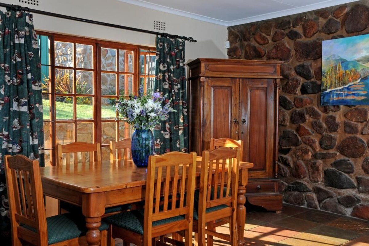 A dining area with a wooden table and chairs, a vase with flowers, a stone wall, and a painting.