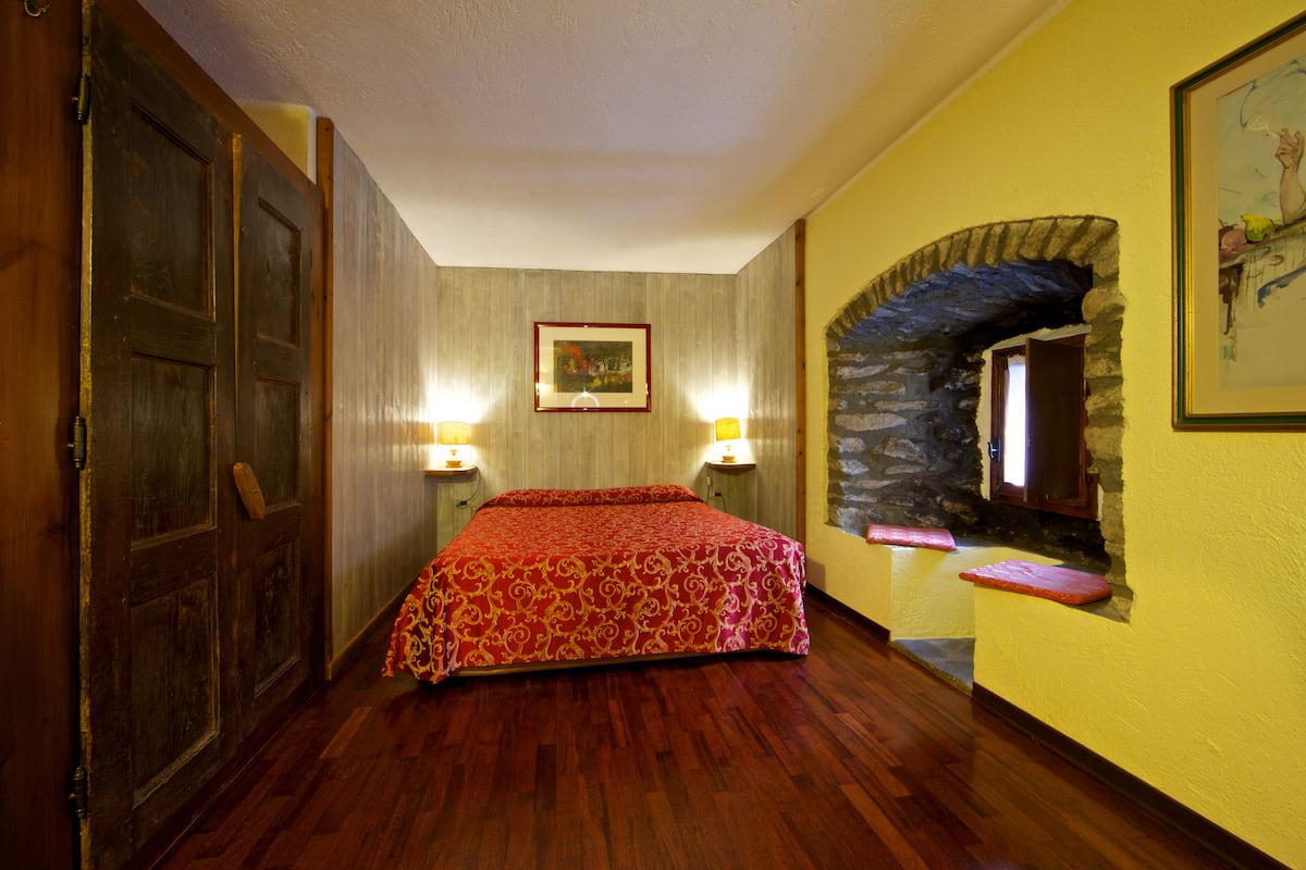 A room with a wooden floor, a bed with a patterned bedspread, two bedside lamps, a framed picture on the wall, and a stone archway.
