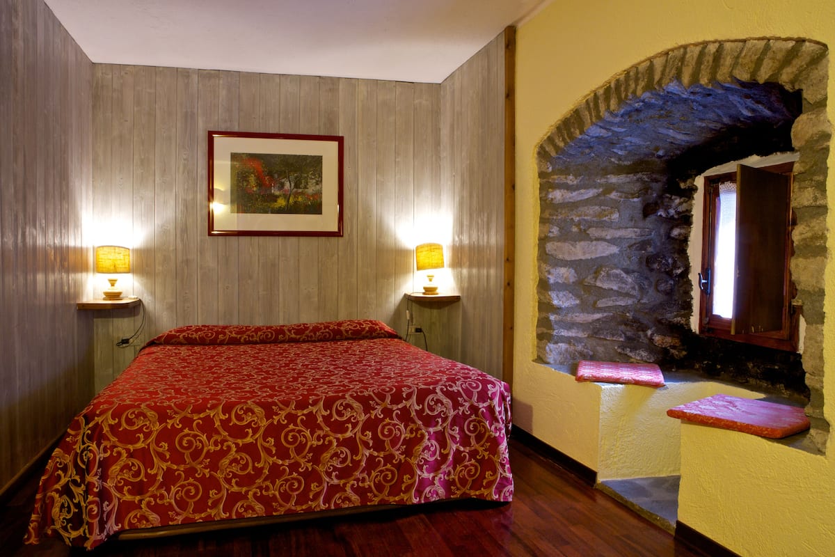 A hotel room with a wooden bed, two wall-mounted lamps, a framed picture, and a stone archway.