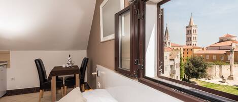 A bedroom with a bed, a table with chairs, a window with a view of buildings, and a door.