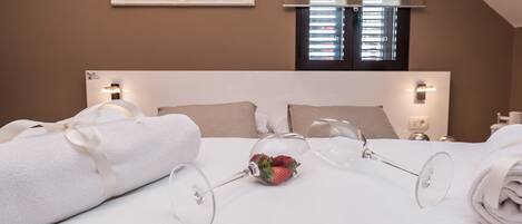 A neatly made bed with a rolled towel and a glass with a strawberry, in a room with a headboard, bedside lamps, and a window with blinds.