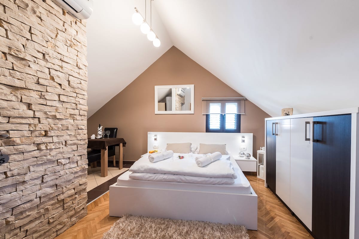 Ein modernes Schlafzimmer mit Steinwand, einem Bett mit weißen Leinentüchern, einem Nachttisch, einem Kleiderschrank und einer Essecke mit Tisch und Stühlen.