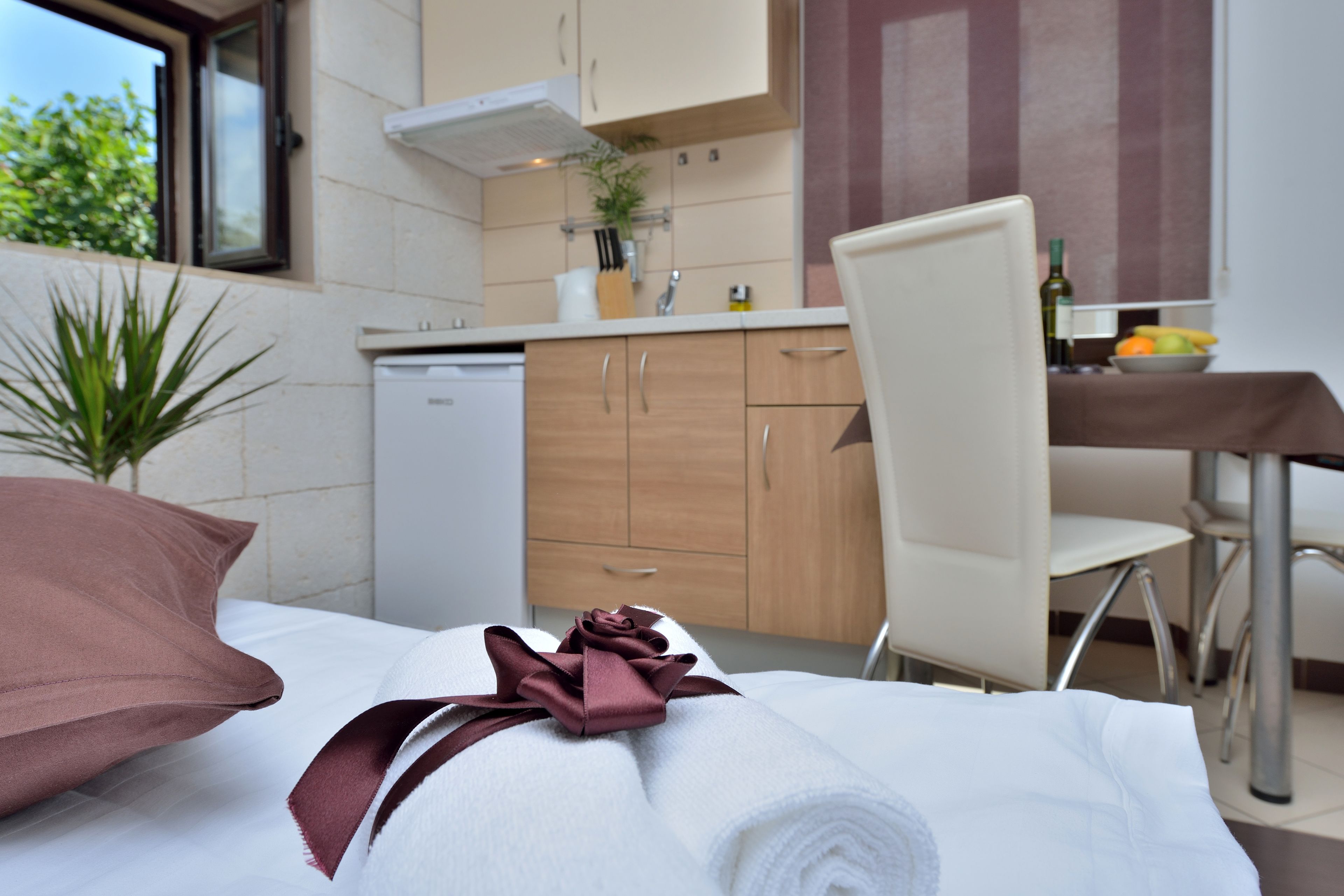 A neatly made bed with a rolled white towel and a brown pillow. A brown ribbon is tied on the towel. A white chair is positioned next to the bed. A small table with a bowl of fruit and a bottle is visible in the background.