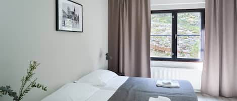 A hotel room with a bed, a framed picture on the wall, a window with curtains, and a vase with a plant.