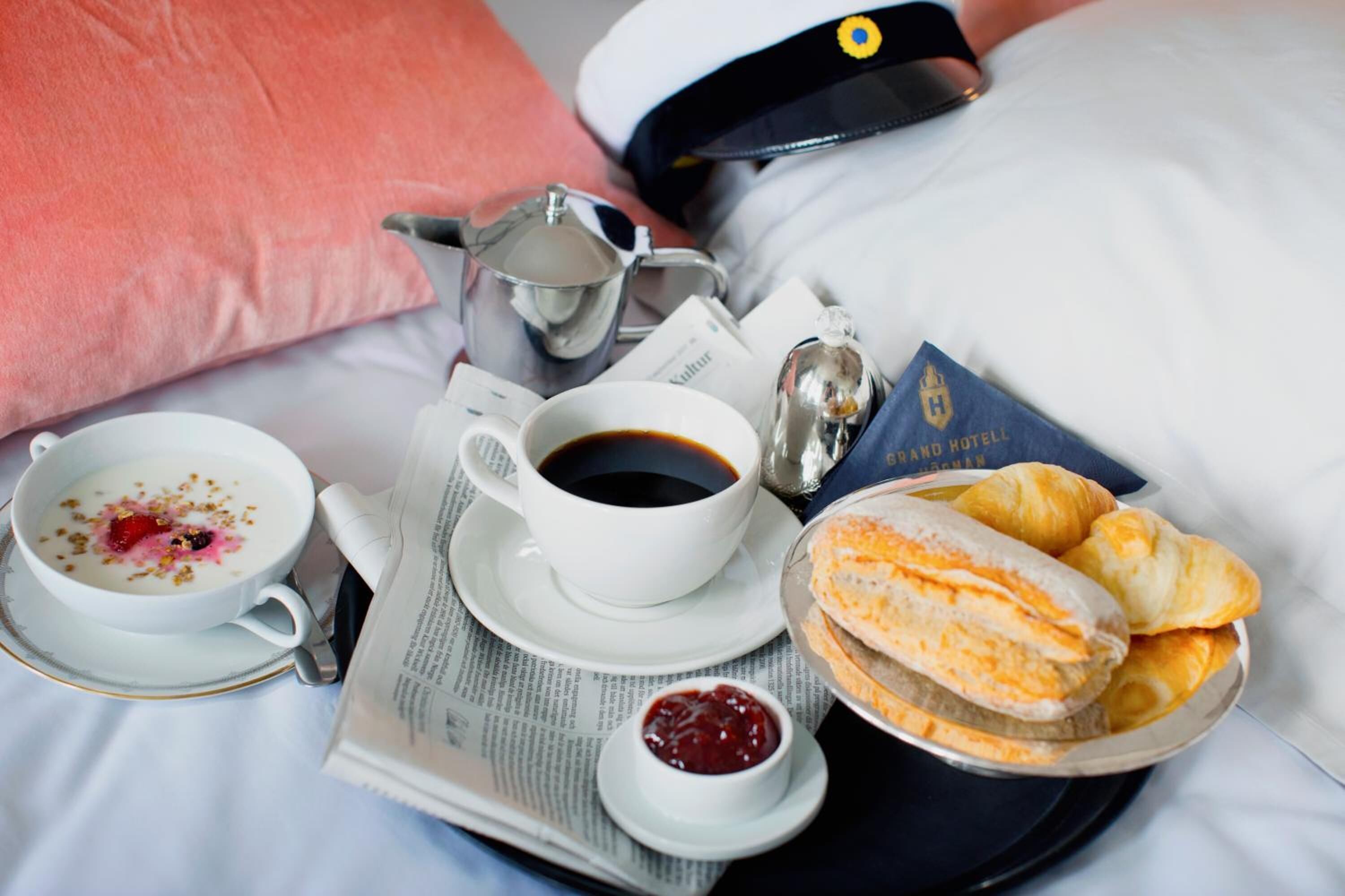 A breakfast tray with a cup of coffee, a bowl of yogurt with fruit, croissants, and a small container of jam on a bed with a pillow and a navy blue cap.