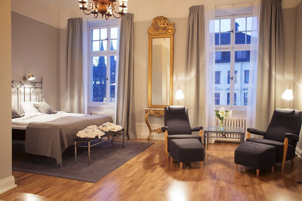 Ein Hotelzimmer mit einem Bett, zwei Sesseln, einem großen Spiegel und Blick auf die Stadt durch große Fenster.