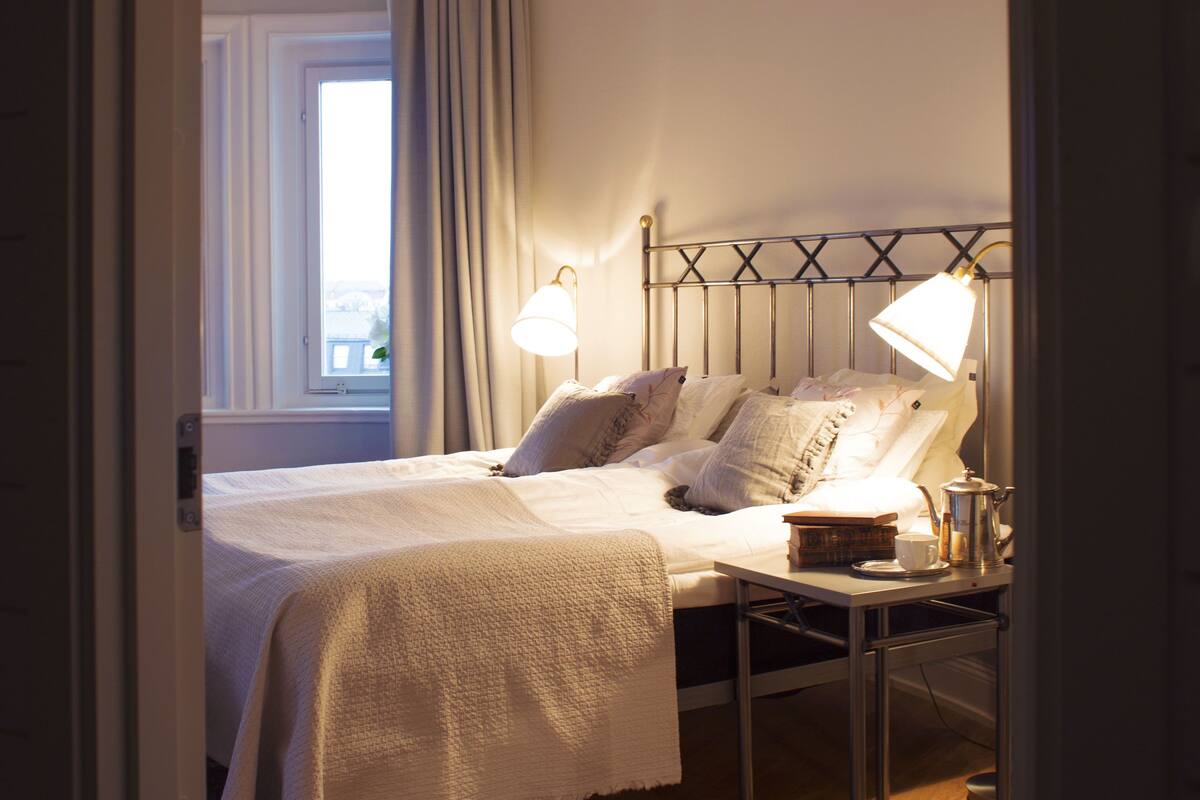 Ein Schlafzimmer mit Bett, Nachttisch, Lampe und einem Fenster mit Vorhängen.