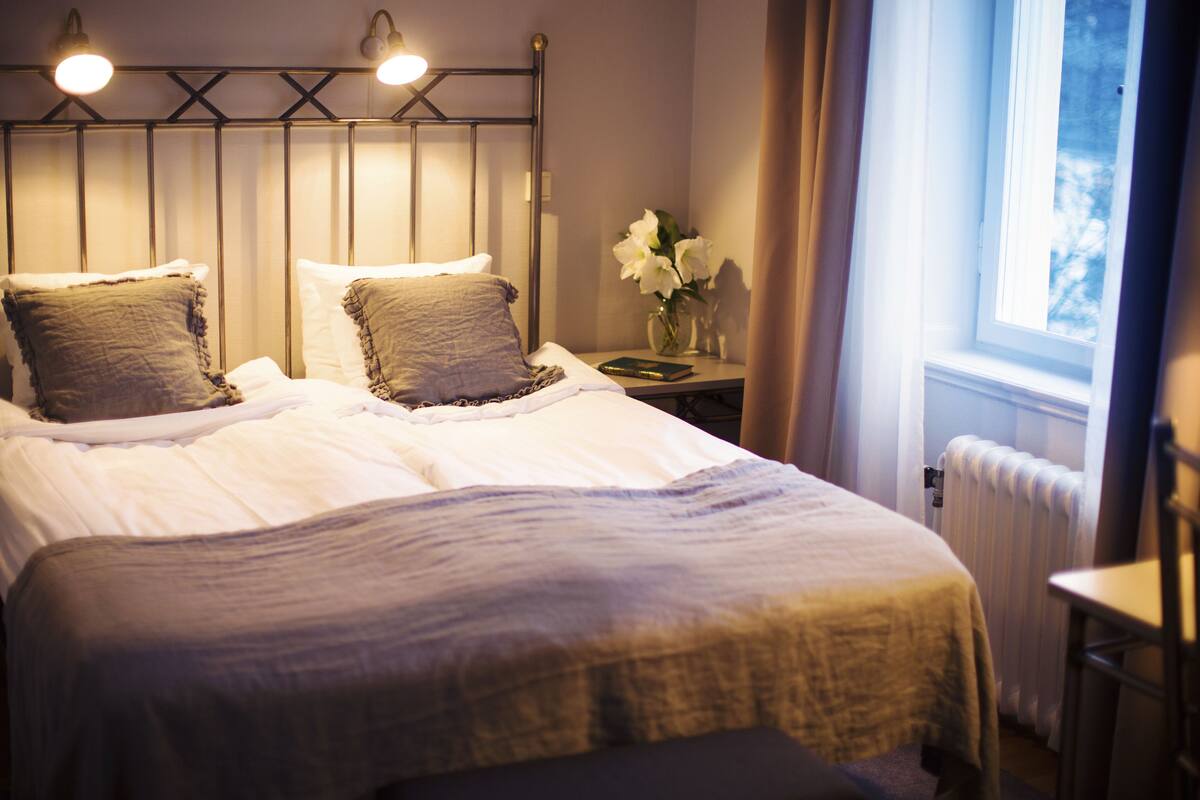 Ein ordentlich bezogenes Bett mit Kopfteil, ein Nachttisch mit Blumenvase und ein Fenster mit Vorhängen.