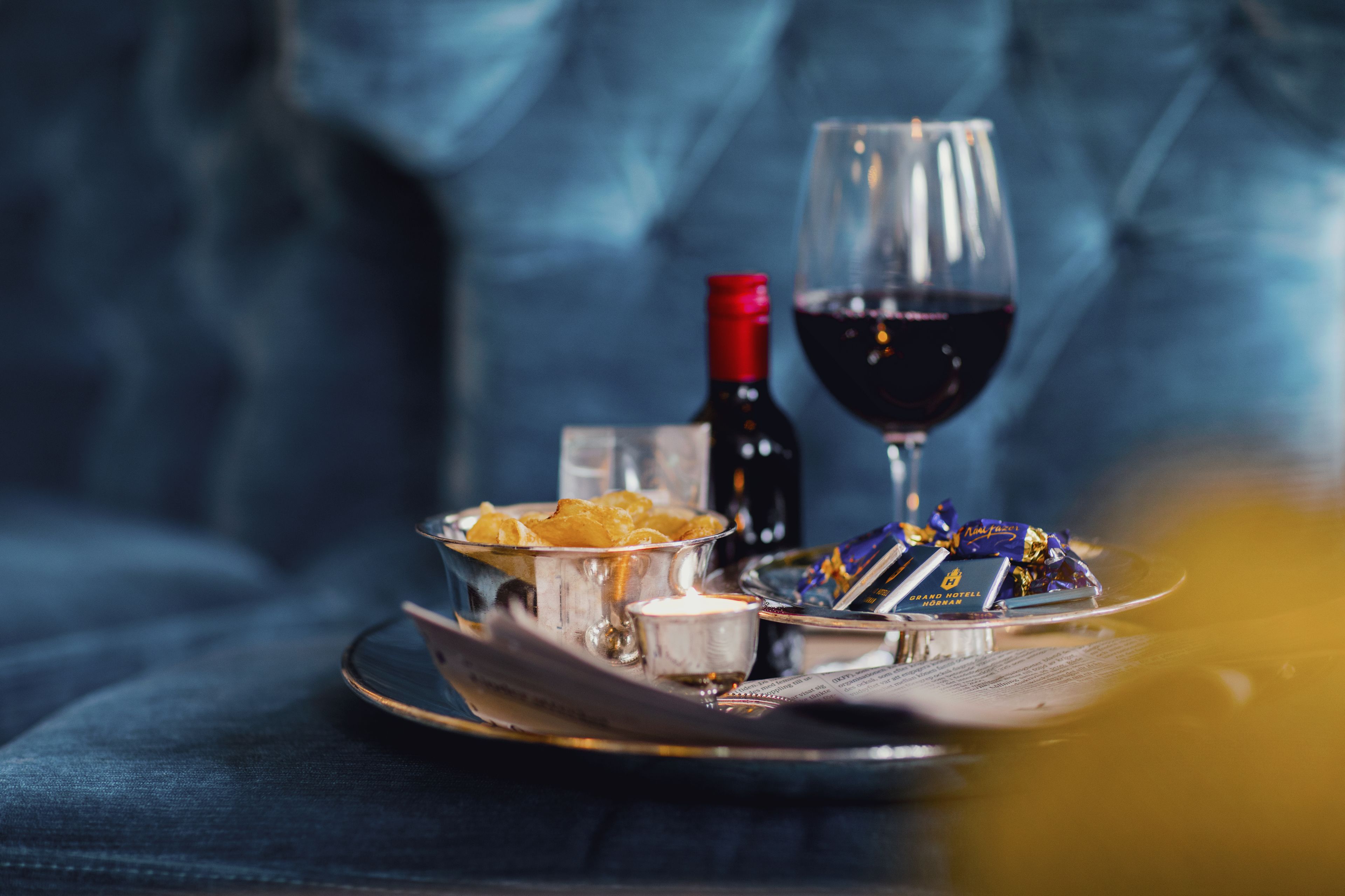 Una bandeja con una botella de vino tinto, una copa de vino tinto, un cuenco pequeño de comida y algunas barras de chocolate sobre una mesa.