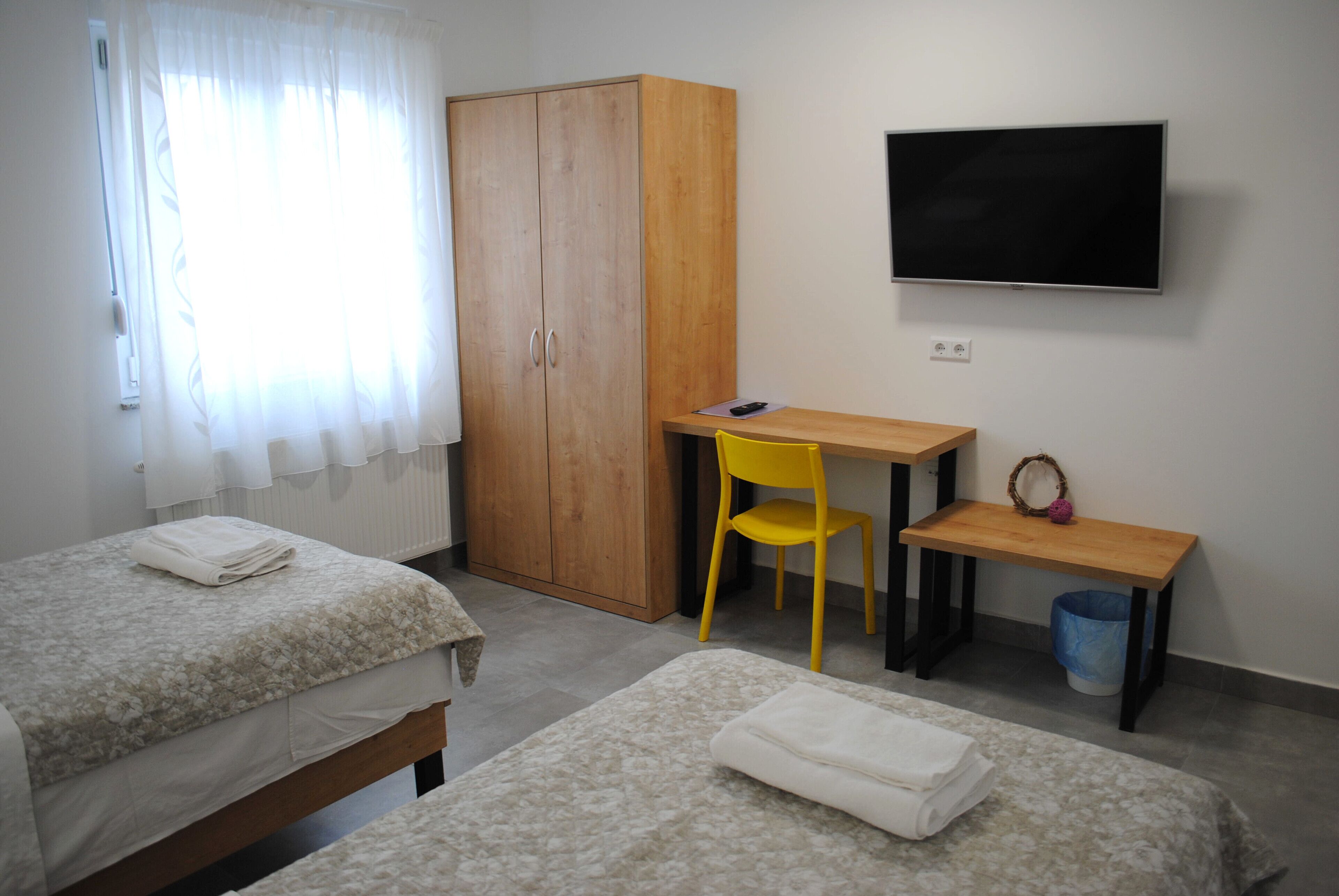 A bedroom with two beds, a wooden wardrobe, a desk with a chair, a television, and a small table.