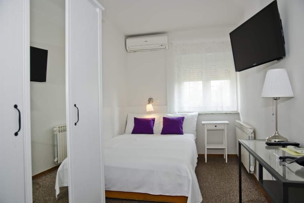 A hotel room with a bed, a desk, a TV, and an air conditioning unit.