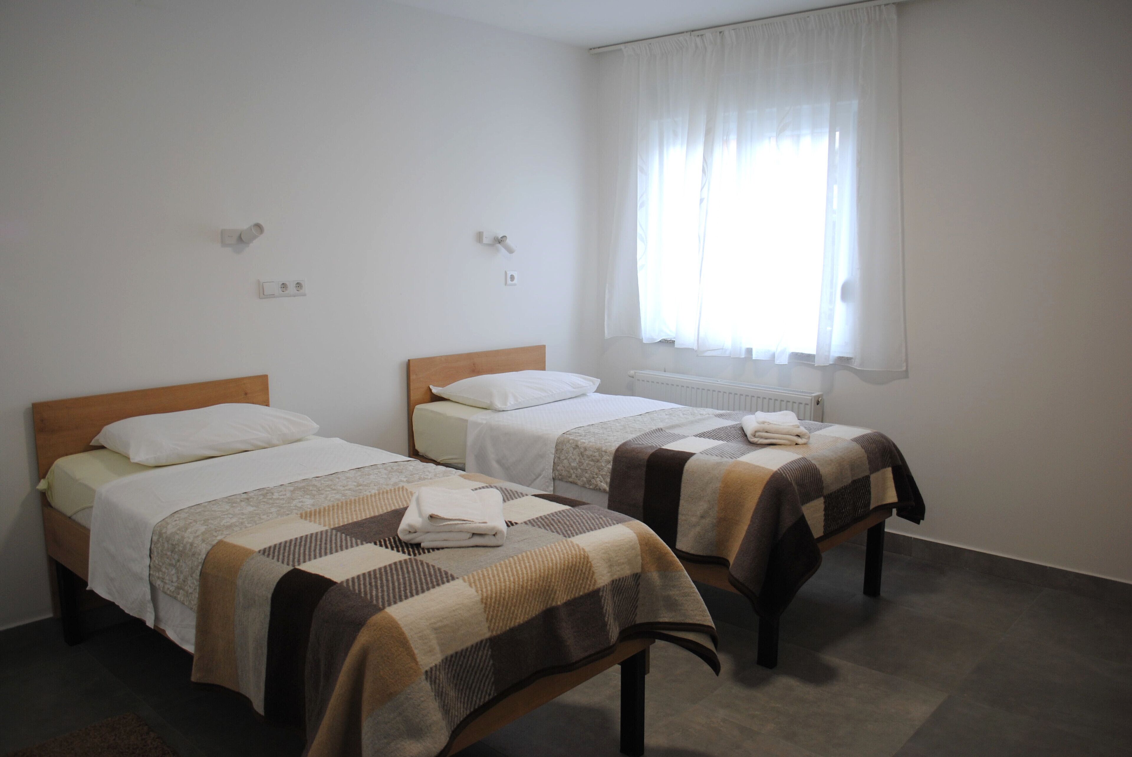 Ein Zimmer mit zwei Einzelbetten, jeweils mit einem Holz-Kopfstück und weißer Bettwäsche. Ein Fenster mit weißen Vorhängen und ein Heizkörper an der Wand sind zu sehen.