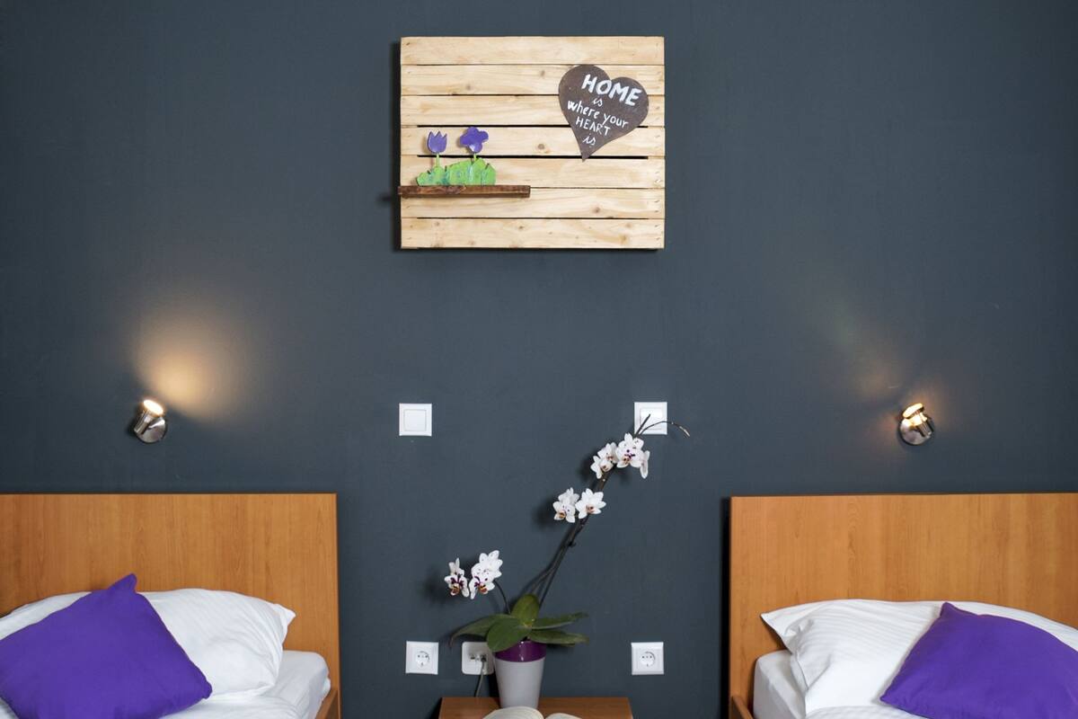A room with two beds, each with a purple pillow and white bedding. There is a small wooden nightstand with a potted orchid and a book between the beds. Above the nightstand, there is a chalkboard sign with the text "HOME is where your HEART is."