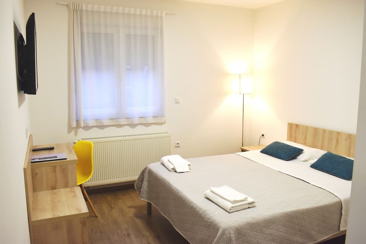 A hotel room with a bed, a desk, a chair, a radiator, a window with curtains, and a wall-mounted TV.