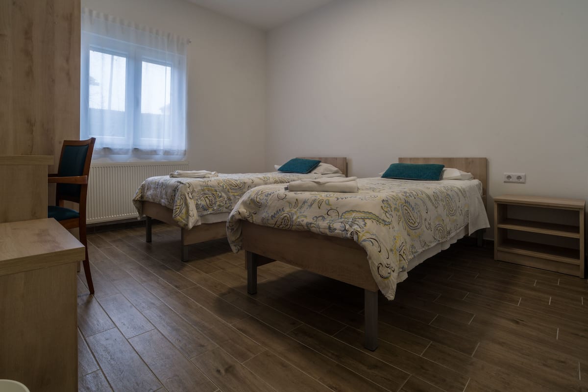 A room with two beds, a chair, a small table, and a window with sheer curtains.