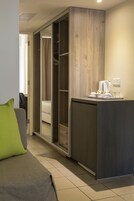 Habitación de hotel moderna con sofá gris, almohada verde, armario de madera y una zona de kitchenette con hervidor y cafetera.
