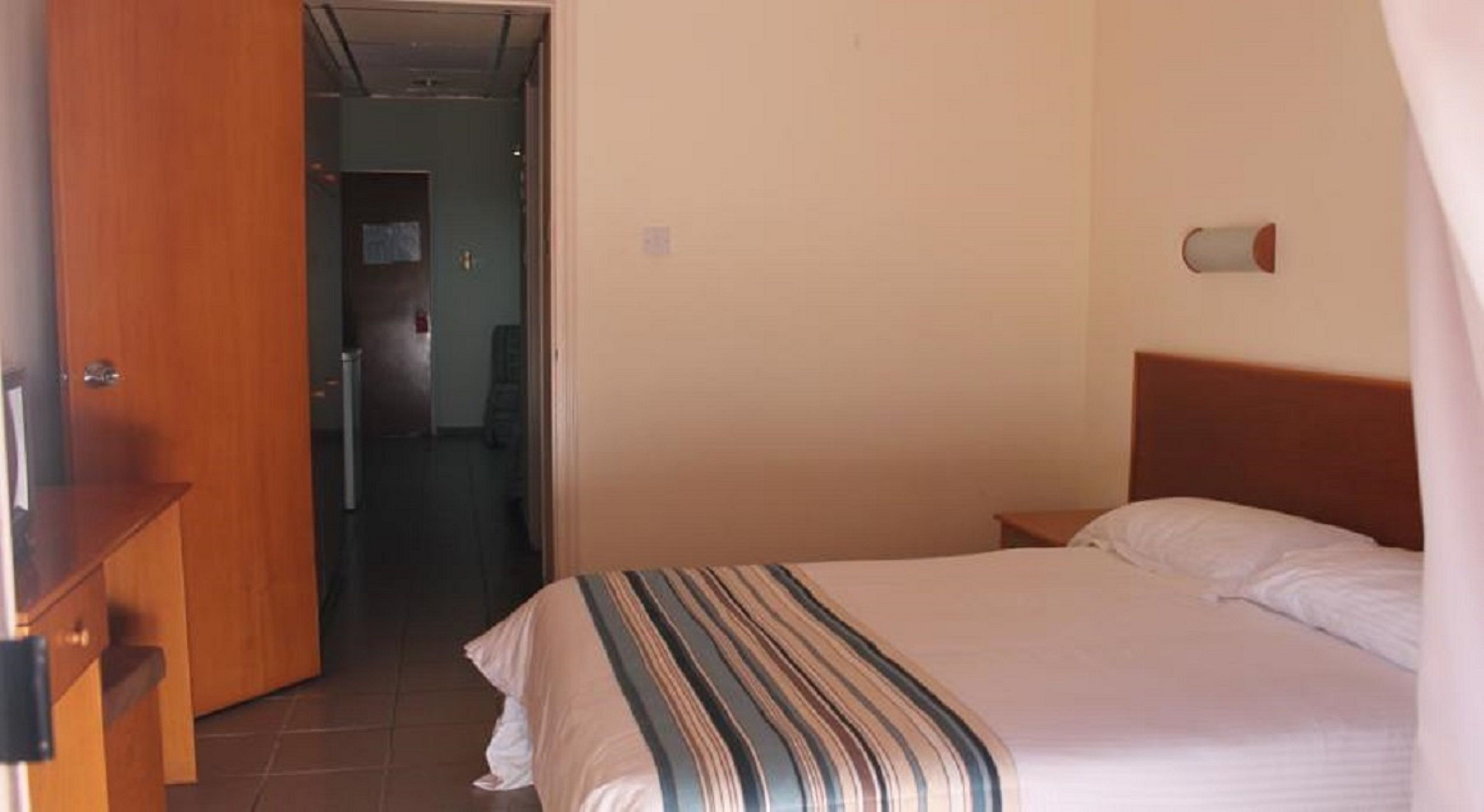 Una habitación de hotel con una cama, una mesita de noche de madera, una puerta que da a otra habitación y un aplique de pared.
