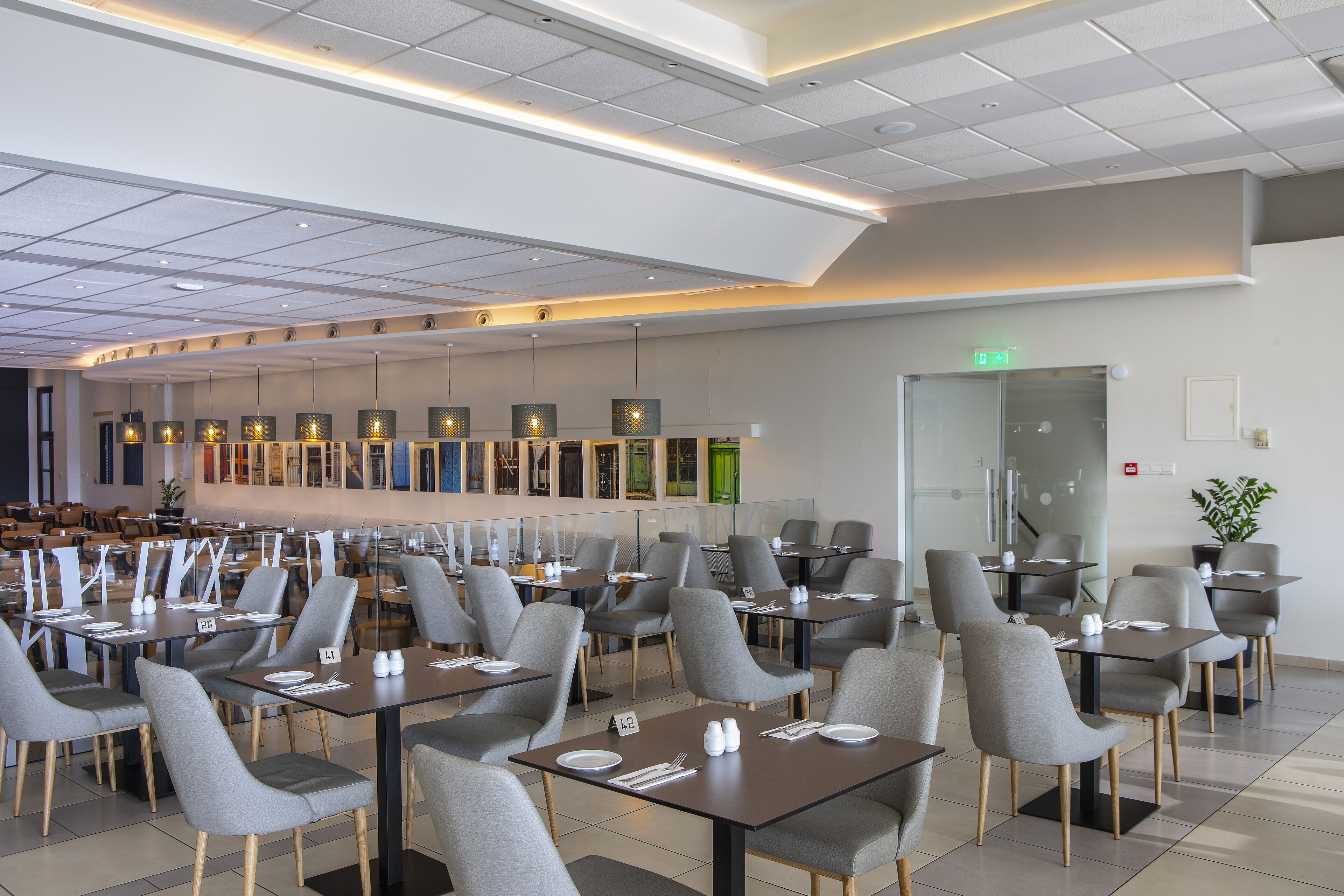 Un restaurant moderne doté de nombreuses tables et chaises, d’un mur de séparation en verre et d’un plafond pourvu d’un éclairage encastré.