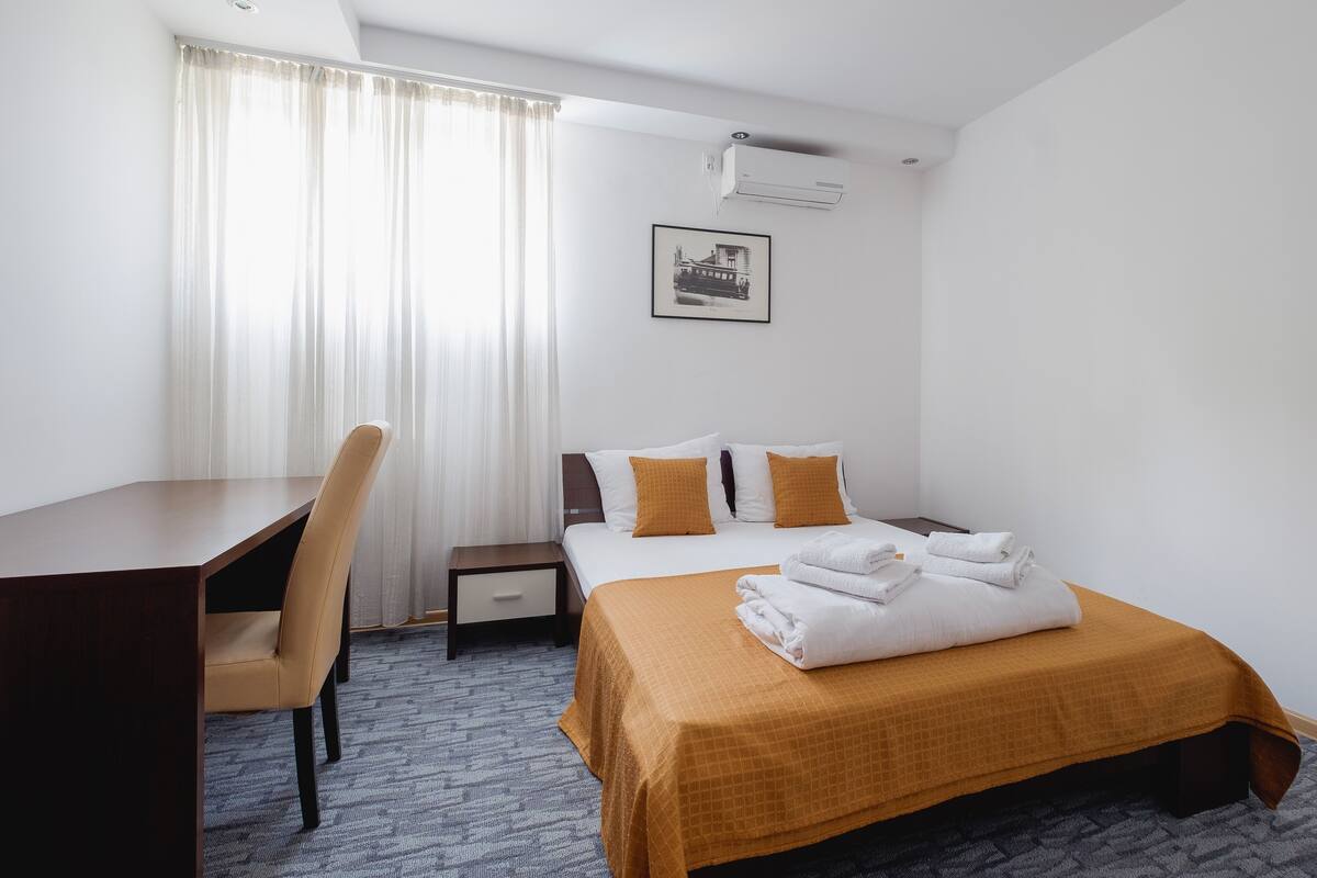 Une chambre d’hôtel comprenant un lit, un bureau, une chaise, une table de chevet et une fenêtre avec des rideaux.