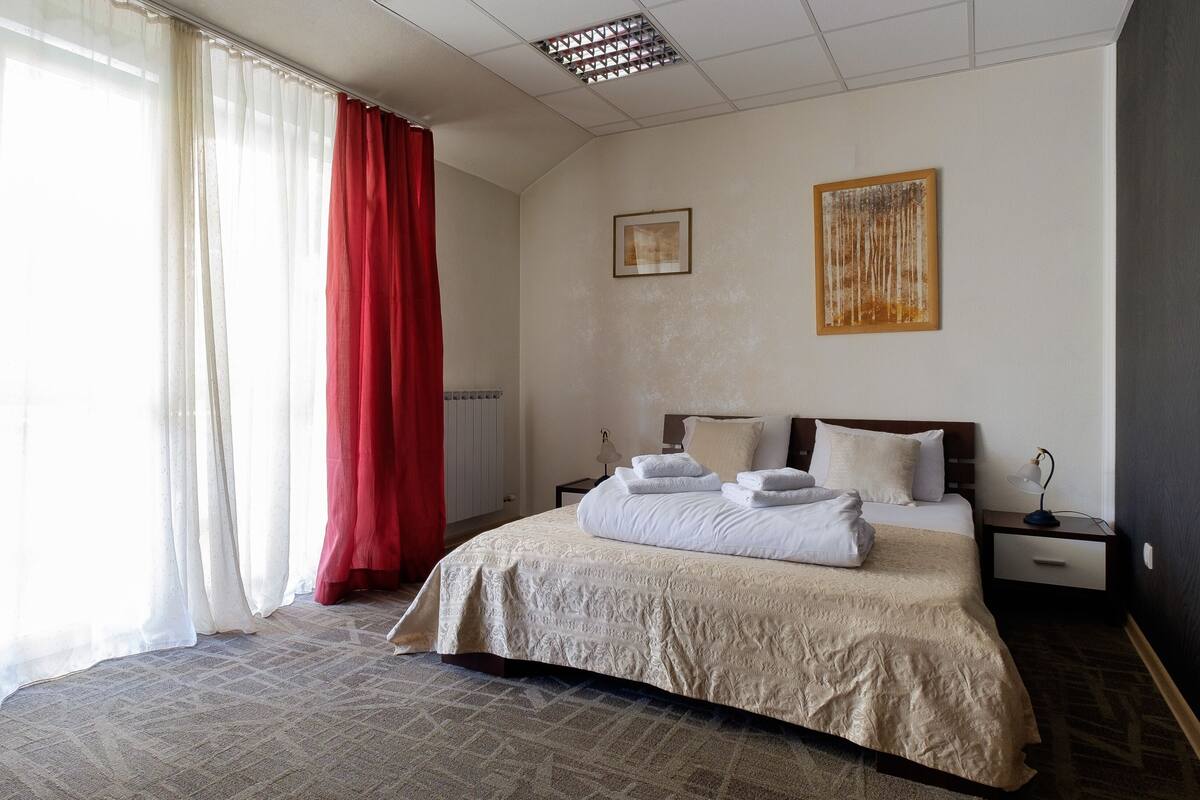 Une chambre d’hôtel avec un grand lit, une table de chevet avec une lampe, un tableau encadré au mur et une fenêtre avec des rideaux rouges.