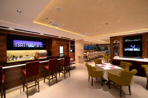A modern bar area with a long counter, red bar stools, and a television screen displaying a bar menu. - Hotel Hedonic (Belgrade)