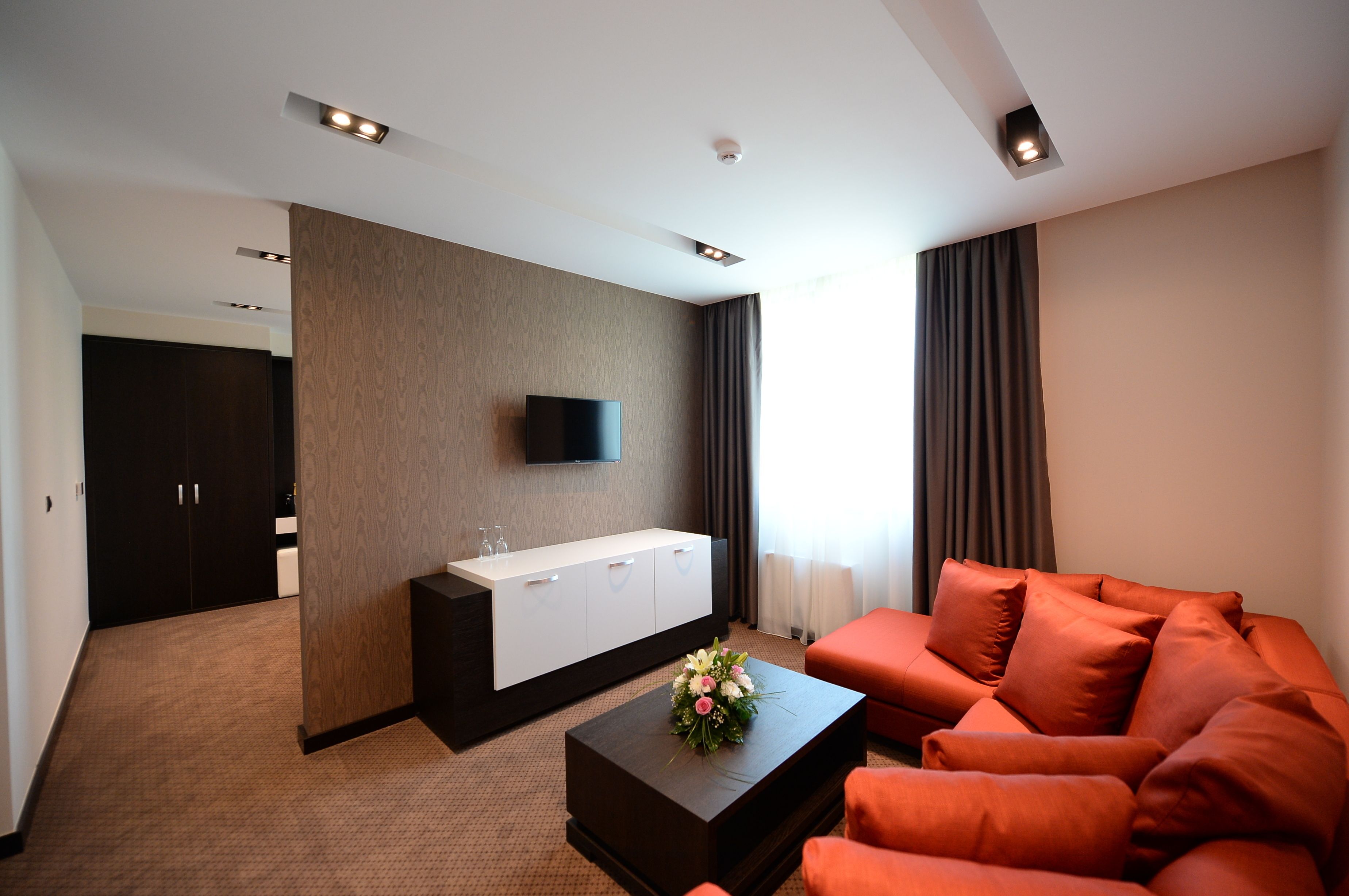 Ein modernes Hotelzimmer mit einer Couch, einem Couchtisch mit floraler Dekoration, einem an der Wand befestigten Fernseher und einem Kleiderschrank.
