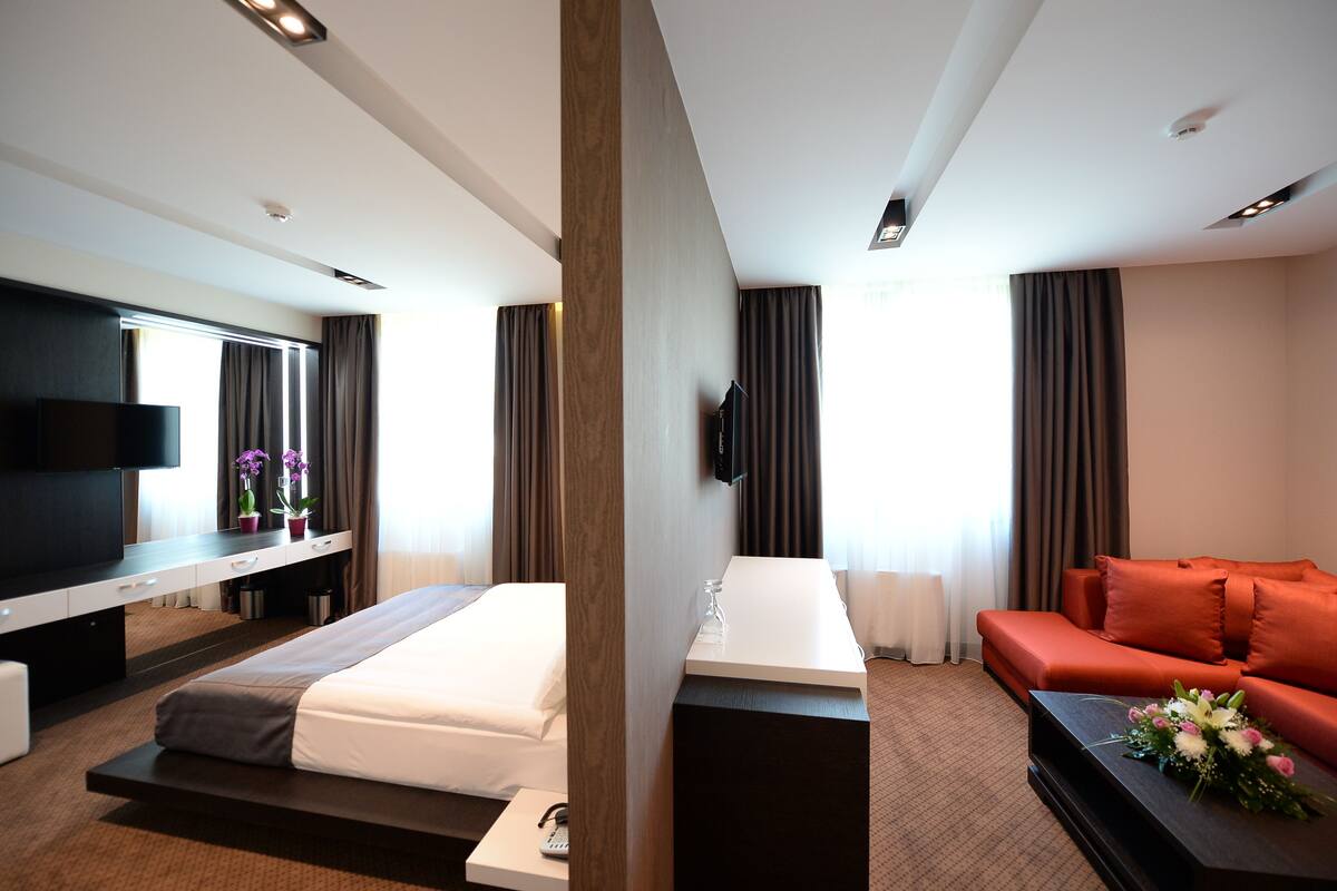 A modern hotel room with a bed, a sofa, a TV, and a desk with a flat-screen TV.