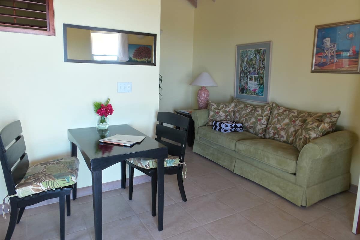 A living room with a green sofa, two black chairs, a table with a vase of flowers, a lamp, and a painting on the wall.
