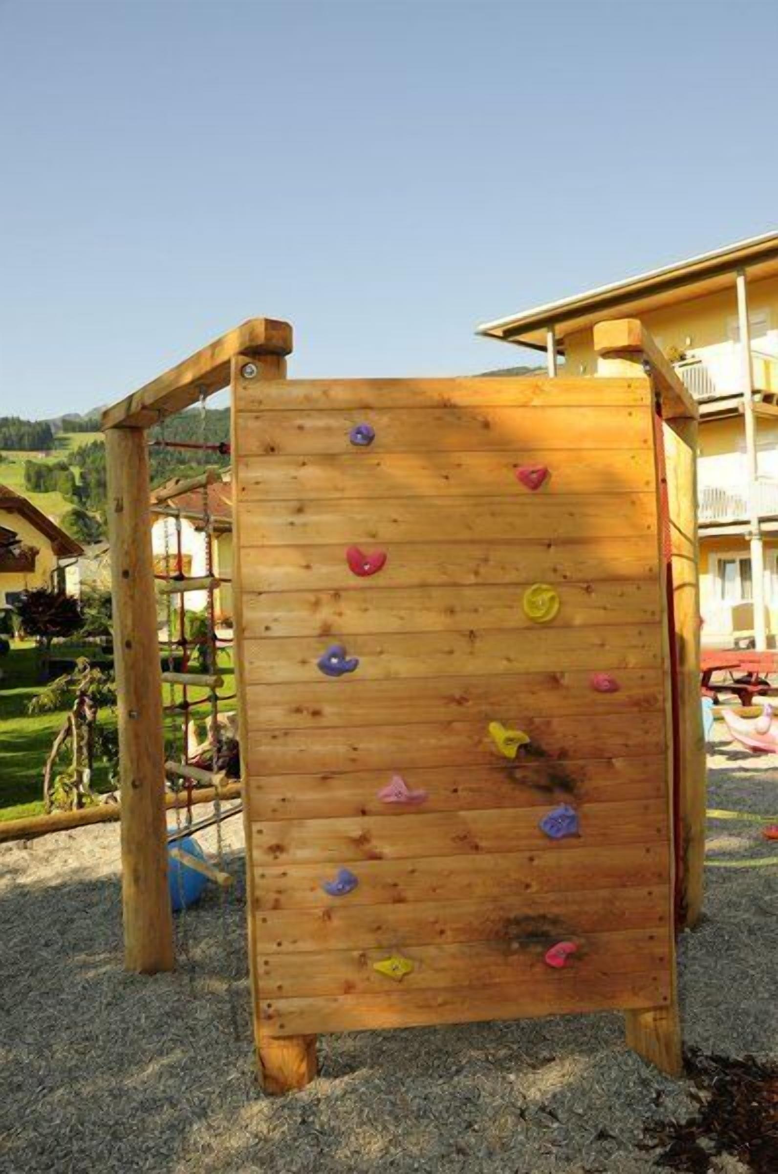 A wooden climbing structure with colorful handholds, set on a gravel surface with a building in the background.