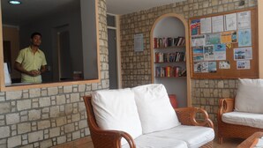Une pièce avec un mur en briques, une bibliothèque en bois, un tableau d’affichage où sont accrochées diverses annonces, et deux canapés en osier.