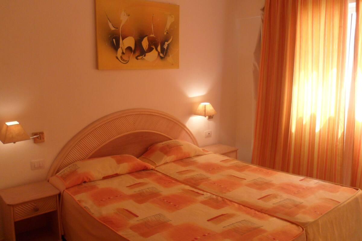 Un dormitorio con una cama, dos mesitas de noche, una lámpara de pared y un cuadro.