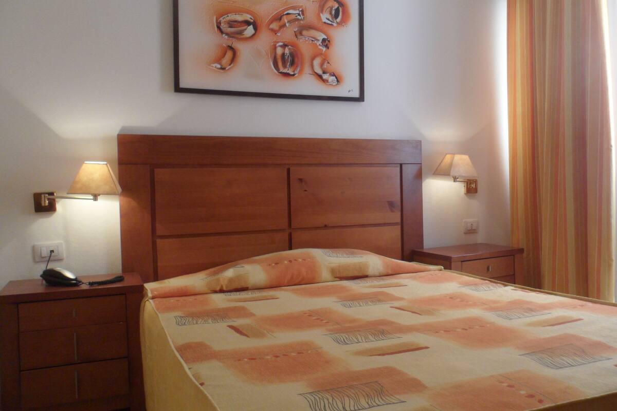 Habitación de hotel con cabecera de madera, dos lámparas de noche, un teléfono y una cama con colcha estampada.