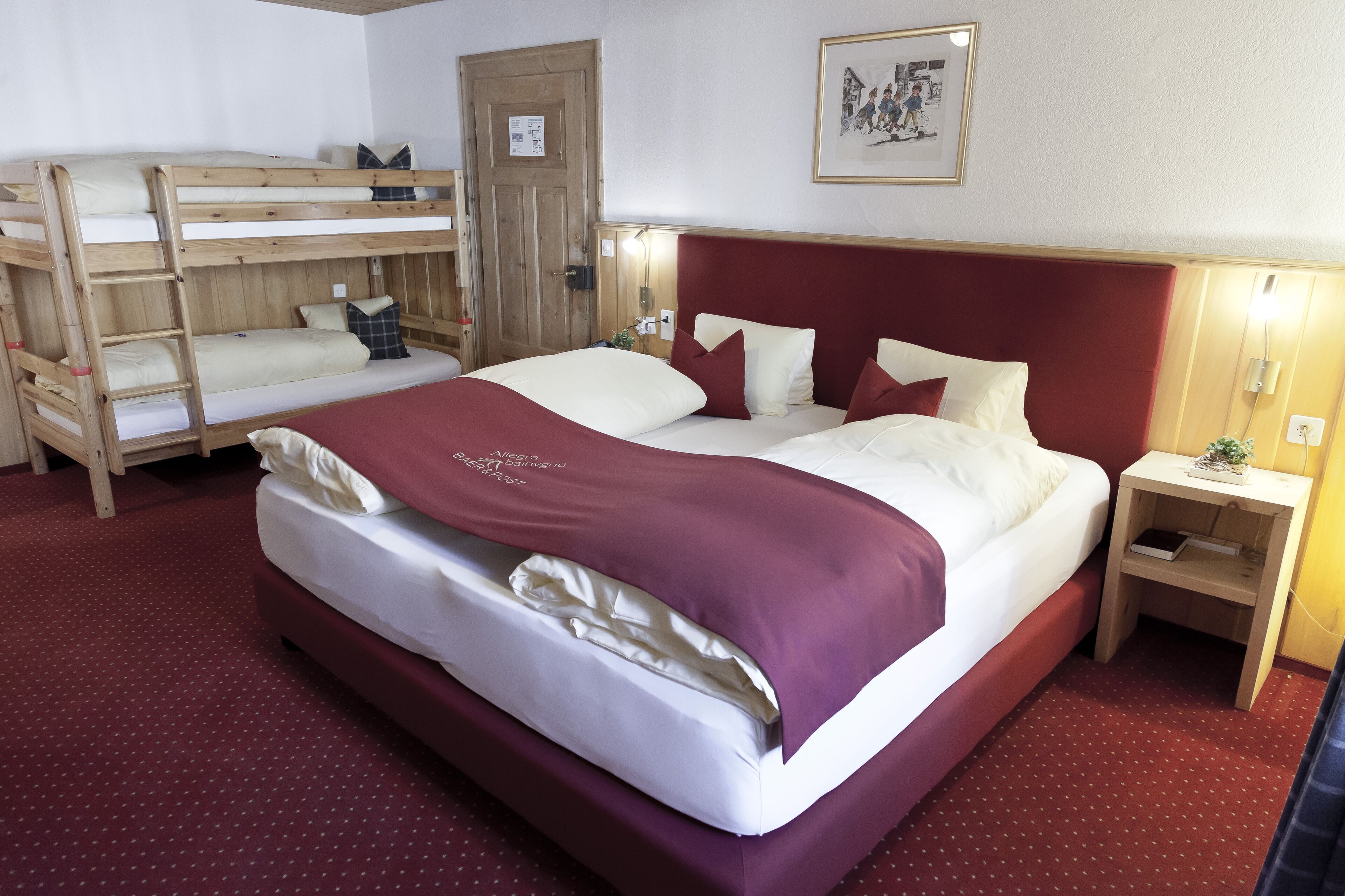 Ein Hotelzimmer mit Etagenbett, einem Einzelbett mit rotem Kopfteil, einem Nachttisch aus Holz mit Lampe und einem gerahmten Bild an der Wand.