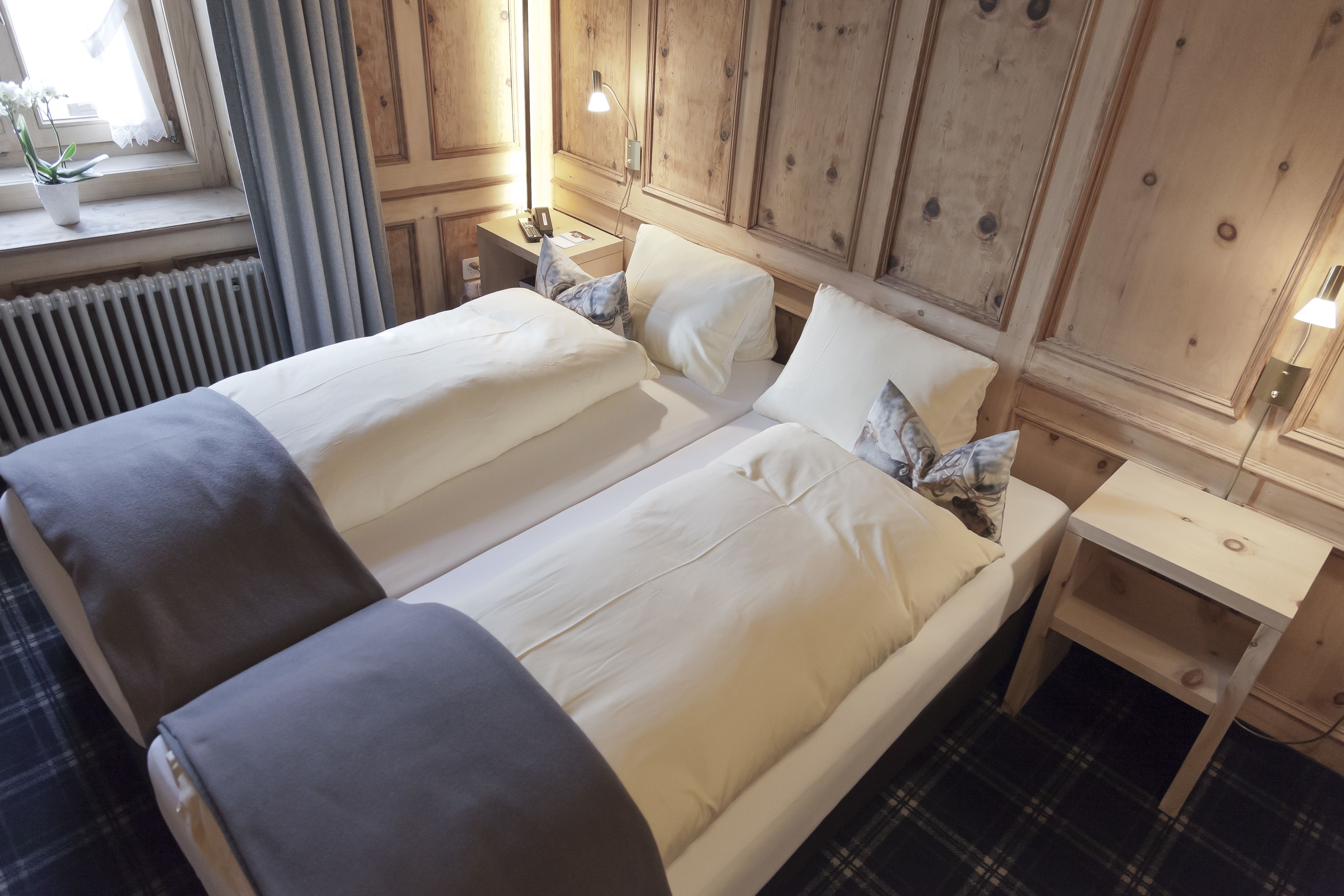 A double bed with white linens and gray throw pillows, a bedside table with a lamp, and a window with curtains.