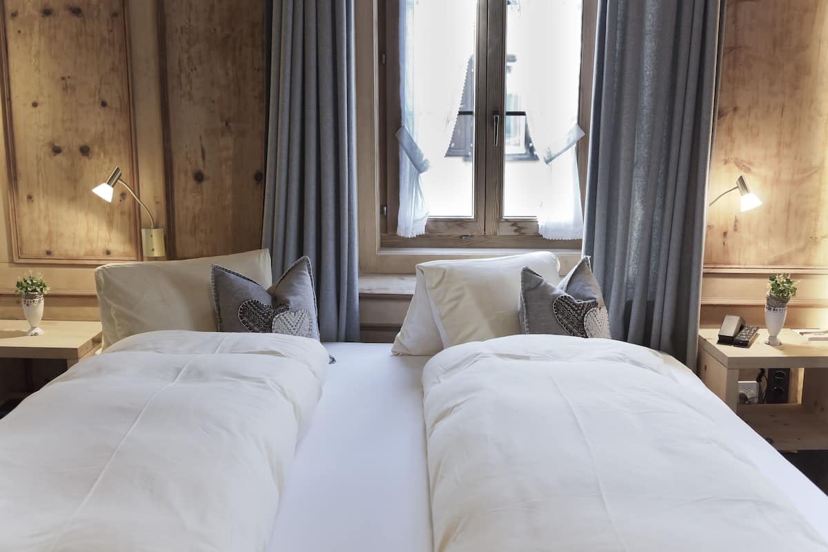 A double bed with white bedding and gray pillows, a bedside table with a lamp, and a window with curtains.