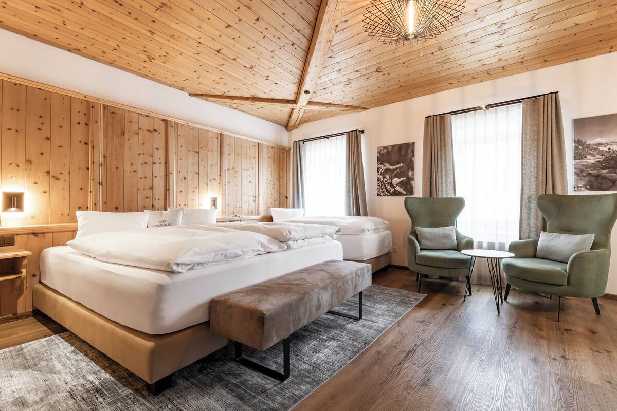 A bedroom with a large bed, two armchairs, a small table, wooden walls, and a ceiling with exposed beams.