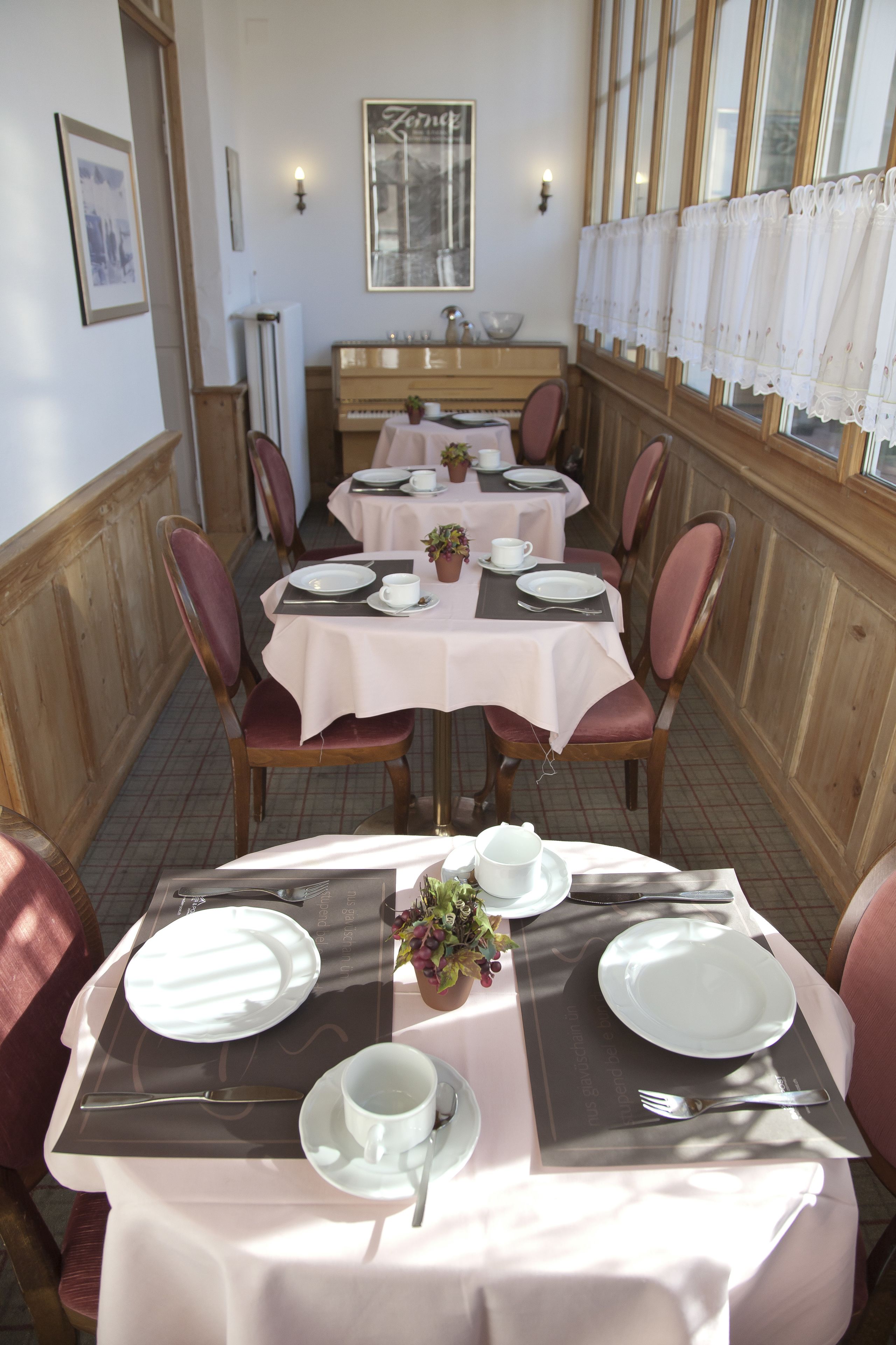 A dining area with tables set for four, each with white tablecloths, plates, cups, and cutlery. There are potted plants on the tables, and the room has wooden paneled walls and large windows with curtains.