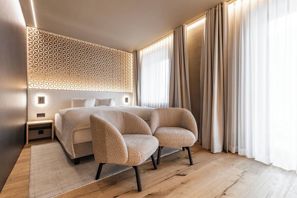 A modern bedroom with a large bed, two upholstered chairs, a patterned wall, and sheer curtains.