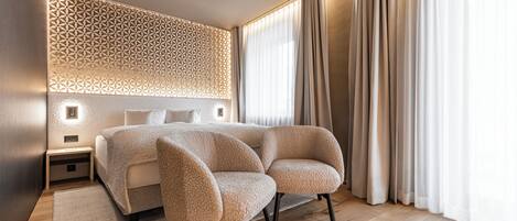 A modern bedroom with a large bed, two upholstered chairs, a patterned wall, and sheer curtains.