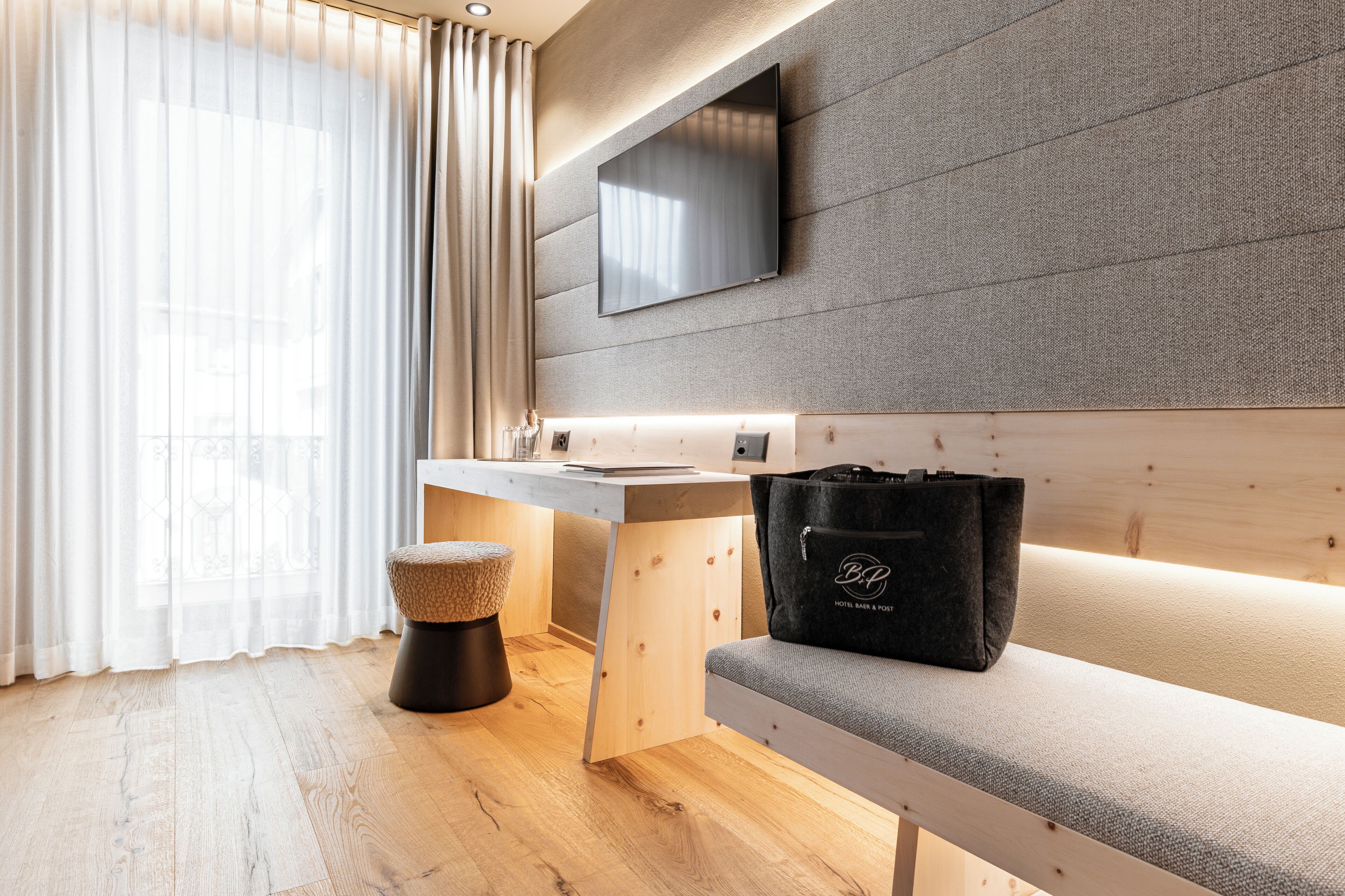 Een moderne hotelkamer met een houten bureau, een bankje, een televisie aan een gestructureerde muur en een raam met dunne gordijnen.