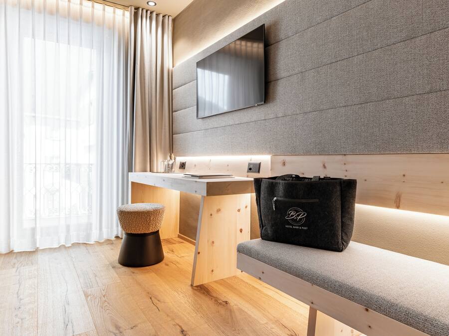 Een moderne hotelkamer met een houten bureau, een bankje, een televisie aan een gestructureerde muur en een raam met dunne gordijnen.