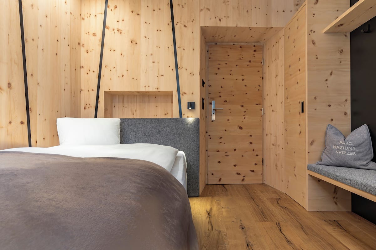 A modern bedroom with a wooden wall, a bed with a grey headboard, and a bench with pillows.